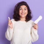 Man sieht eine junge Dame mit einem Tampon und einer Binde in der Hand vor einem lila Hintergrund
