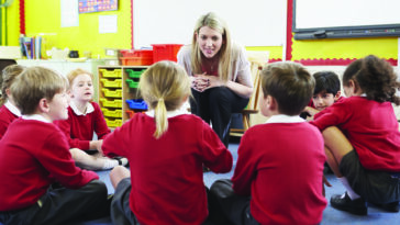 Auf dem Bild ist eine Gruppe von Kindergartenkindern zu sehen, die am Boden im Kreis sitzen. Ihnen gegenüber beugt sich eine Kindergartenpädagogin lächelnd zu ihnen herunter.