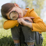 Forscherinnentag Seewinkel 2023 ein Mädchen sitzt in der Wiese und hält ein Fernglas