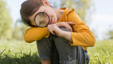 Forscherinnentag Seewinkel 2023 ein Mädchen sitzt in der Wiese und hält ein Fernglas