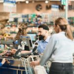 Der Diskonter Lidl senkt die Preise. Am Bild sieht man einen Supermarktkassierer mit Bart, einen Buben links und eine Frau rechts vom Förderband.