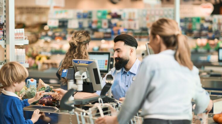 Der Diskonter Lidl senkt die Preise. Am Bild sieht man einen Supermarktkassierer mit Bart, einen Buben links und eine Frau rechts vom Förderband.