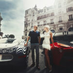 Reiche gönnen sich trotz Krise einen luxuriösen Lebensstil. Am Bild sieht man ein Pärchen zwischen zwei Luxusautos posieren.