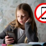 Mädchen sitzt am Handy während neben ihr Schulbücher geöffnet sind. Daneben ist ein großes "Handy verboten"-Zeichen. handyverbot-schule-oesterreich