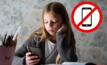 Mädchen sitzt am Handy während neben ihr Schulbücher geöffnet sind. Daneben ist ein großes "Handy verboten"-Zeichen. handyverbot-schule-oesterreich
