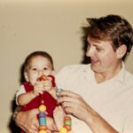 Vater hält Baby im Arm, das mit einer bunten Spielzeugkette spielt.