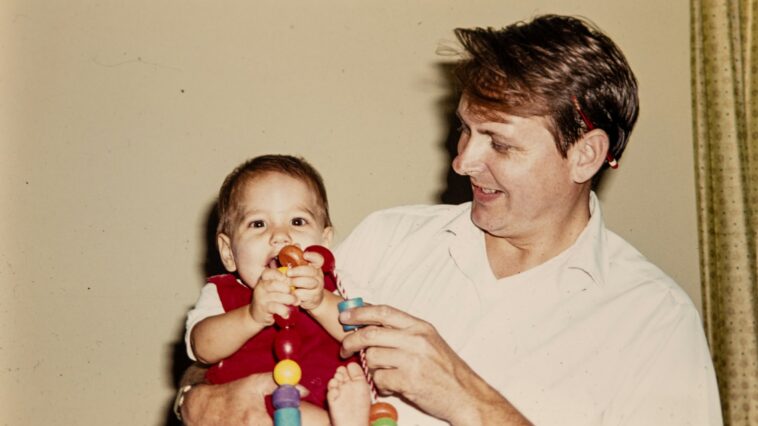 Vater hält Baby im Arm, das mit einer bunten Spielzeugkette spielt.