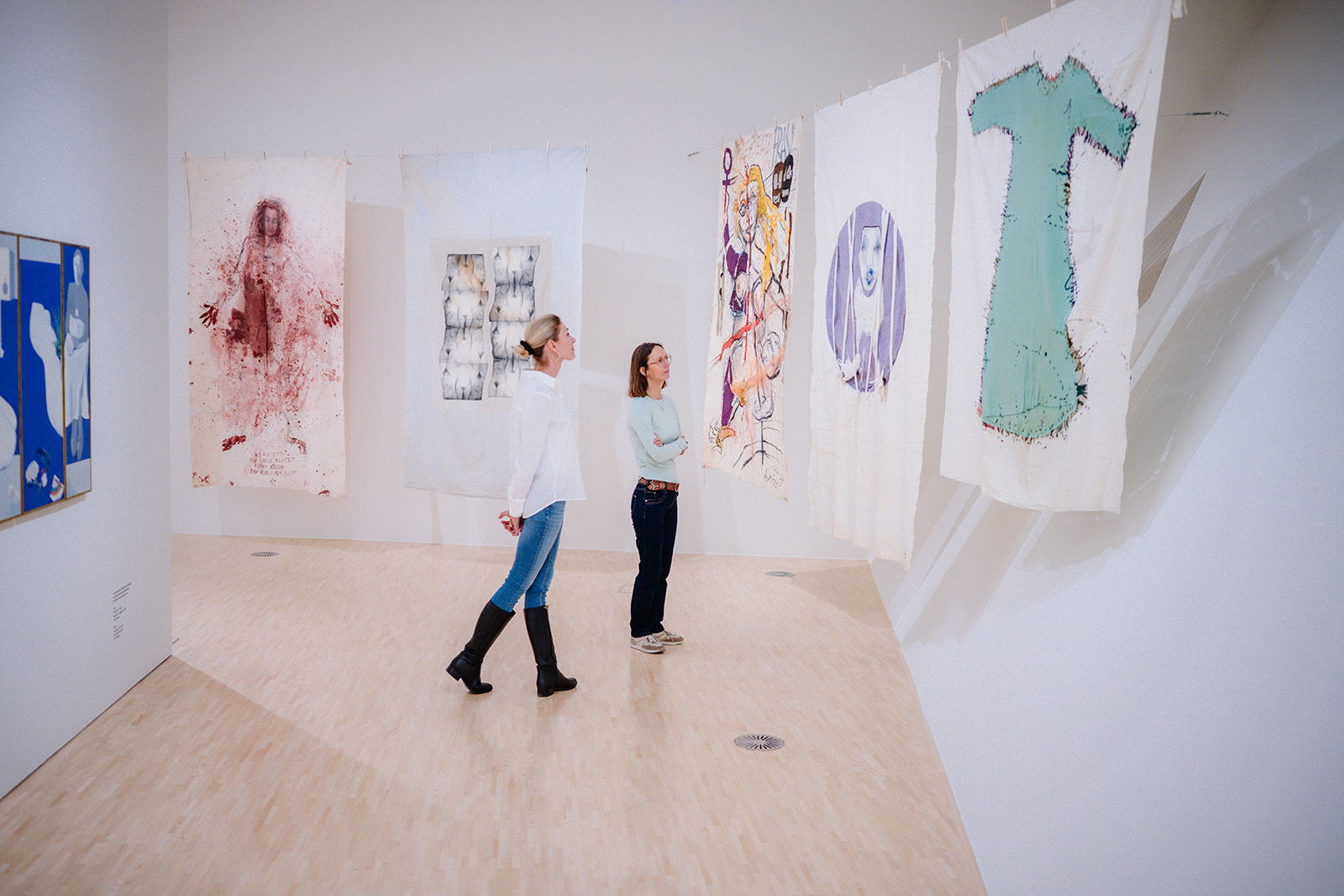 Das Foto zeigt die Ausstellung in der Landesgalerie Krems. Zu sehen sind zwei Frauen, die Leintücher mit verschiedenen Designs zu den Themen Geburt, Menstruation, Sexualität und Tod begutachten.